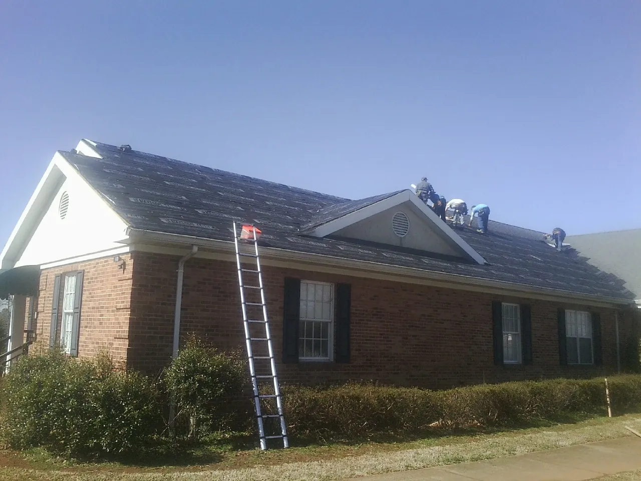 Crew tearing shingles off a brick ranch roof with a ladder set — Emergency Roof Repair work by our crew in Center Moriches