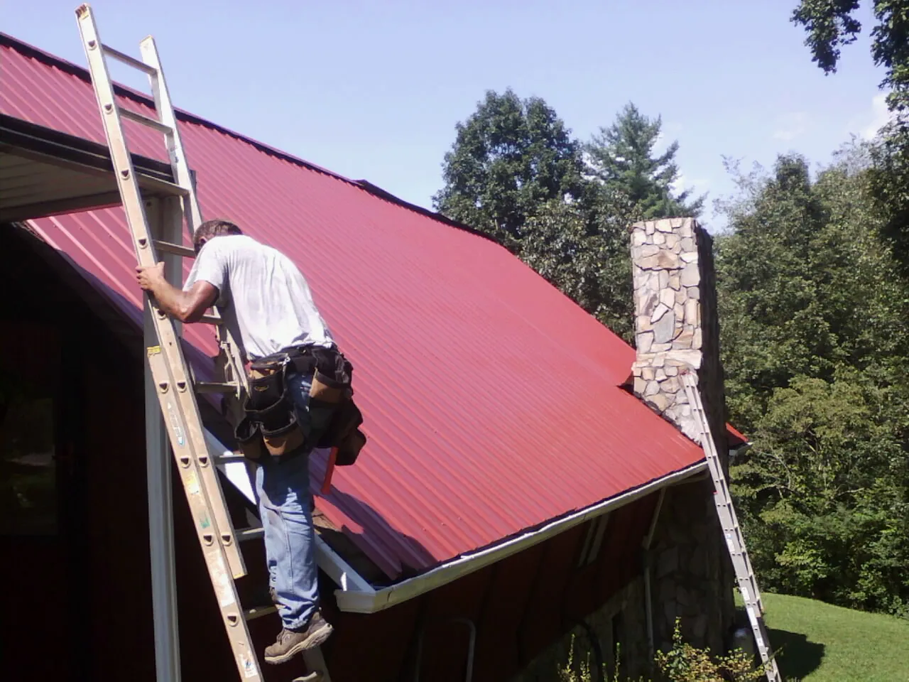 Roofer on a ladder beside a red metal roof and stone chimney — Soffit & Fascia Repair by Roofing Brothers in Center Moriches