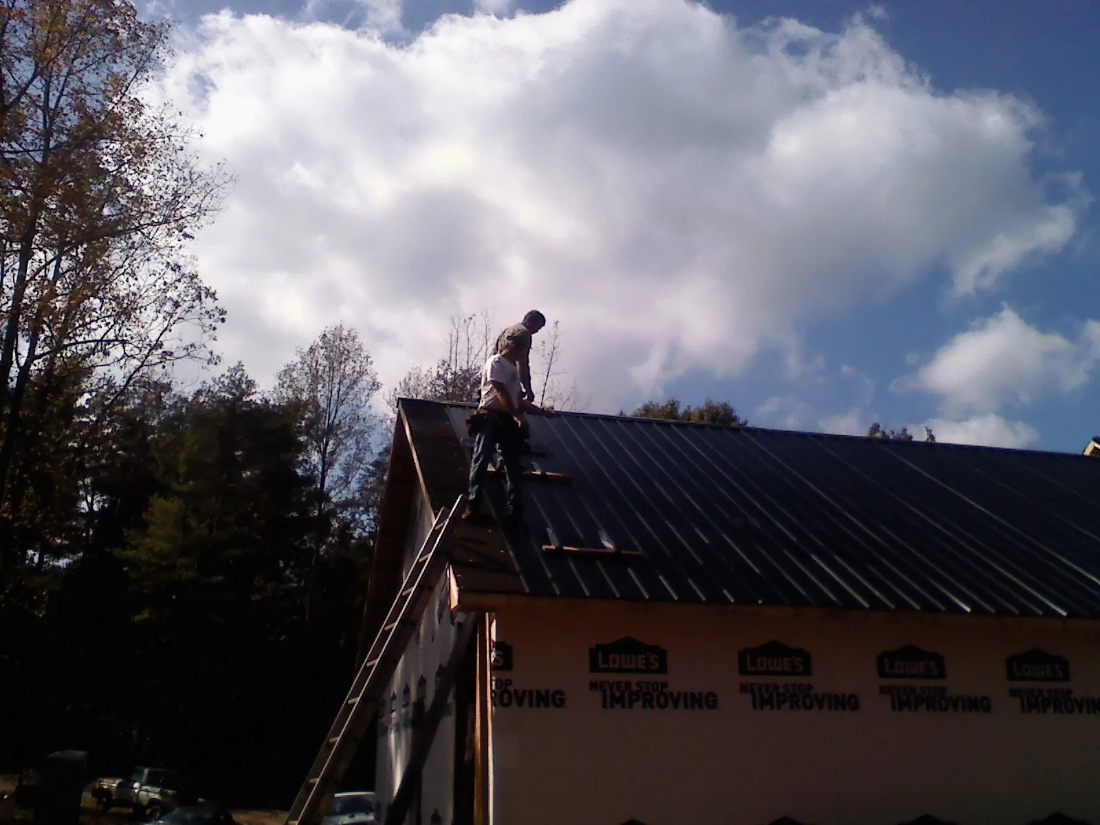 Roofer fastening dark metal panels high against a cloudy sky, from a Roof Inspection job in Center Moriches